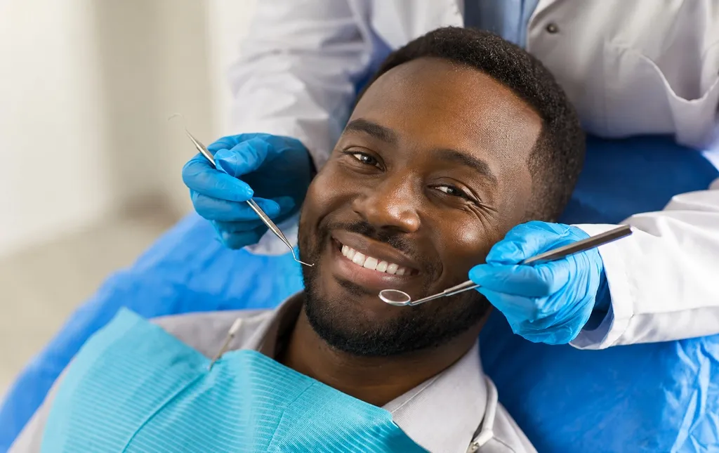 A smiling Black man reclining in a dental chair while a dentist in blue gloves holds dental mirrors for a checkup